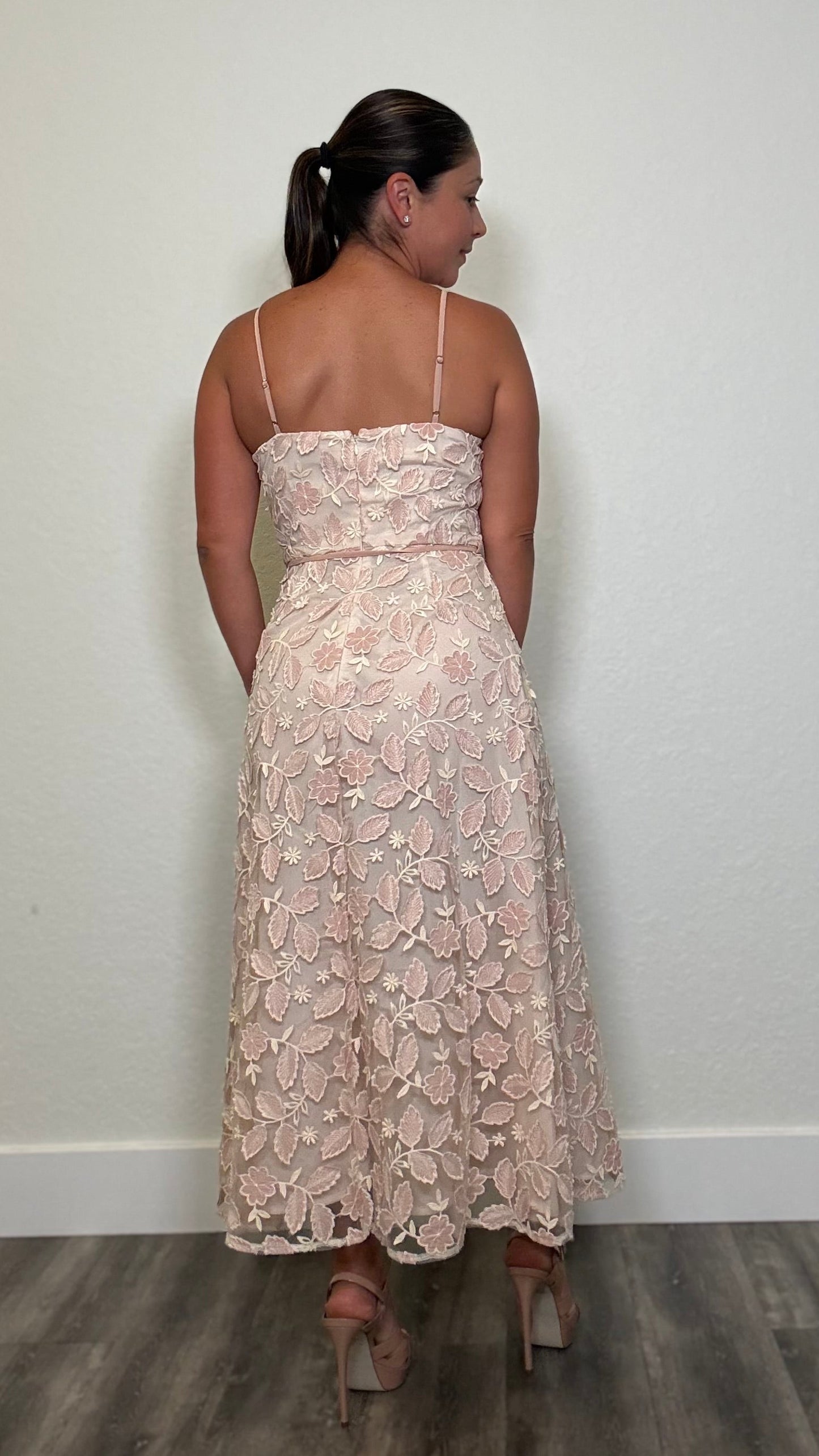 A woman viewed from behind wearing a sleeveless, floor-length lace dress in a pale blush pink color.
