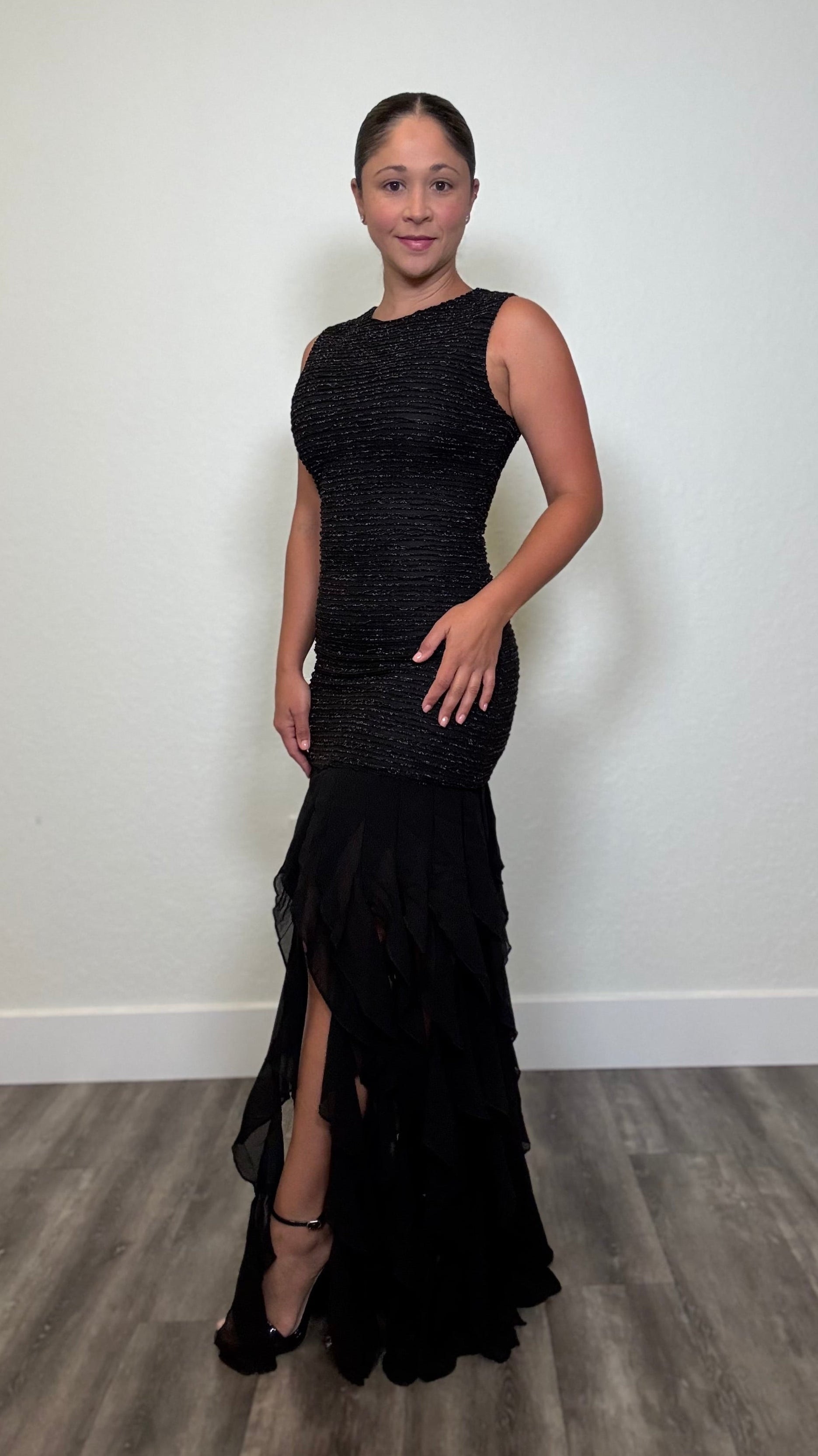 A woman modeling a sleeveless black mermaid-style evening gown with a textured bodice, fitted skirt, and layered ruffle hem